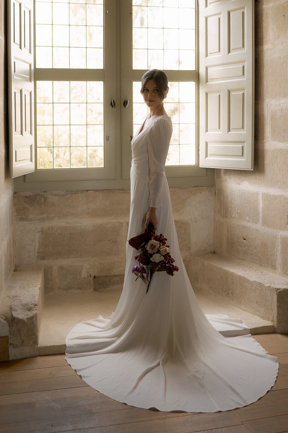 Mariée en robe longue à traîne devant une fenêtre, maquillage naturel longue tenue